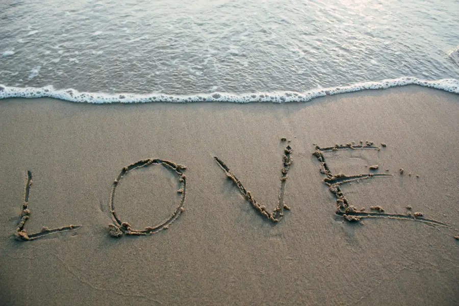 The word "Love" written in the sand at the beach with ocean waves, representing the question Why Can't I Find Love