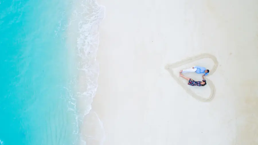 A couple lying inside a large heart drawn in the white sand of a tropical beach – the perfect setting for a surprise romantic getaway.